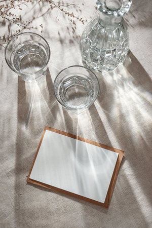 Blank paper card with copy space, glasses and bottle with water or alcohol on a neutral beige table, aesthetic sunlight shadows, invitation or business brand templateの写真素材
