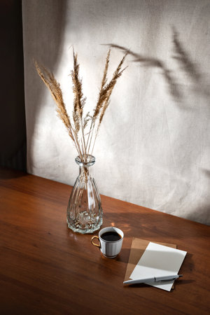 Aesthetic summer interior still life with cop of coffee, paper cards and dry grass in glass vase on a wooden table, floral sunlight shadows on a wall, morning business conceptの写真素材
