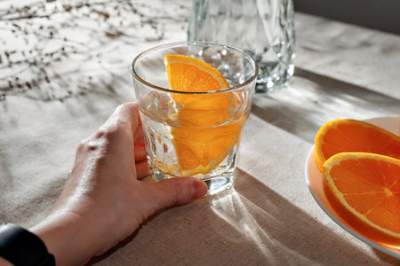 Hand holding glass with orange infused water, bottle, sliced oranges on a beige table with aesthetic sunlight shadows, vegan, healthy morning drink conceptの写真素材