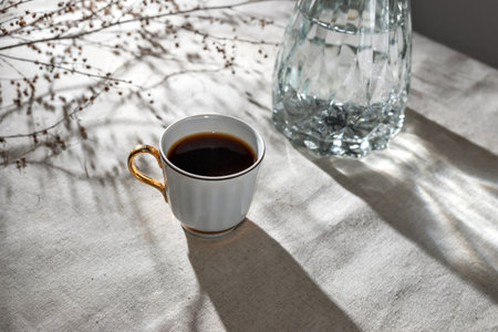 Aesthetic minimalist morning concept, cup of coffee, glass vase and floral sunlight shadows on a neutral beige table clothの写真素材