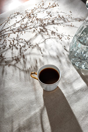 Elegant minimalist morning concept, cup of coffee, glass vase and aesthetic sunlight shadows on a neutral beige table cloth, top view, flat layの写真素材