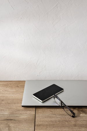 Elegant minimalist business background, home office workspace with laptop, mobile phone and glasses on a beige wooden table, white wall copy spaceの写真素材