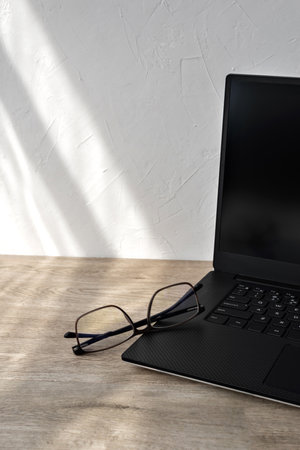 Minimalist business branding concept, laptop and glasses on a wooden table, geometric sunlight shadows on white wall background, copy spaceの写真素材