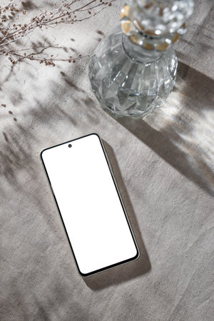 Mobile phone with blank screen mock up, bottle with water or alcohol on a neutral beige linen table cloth background, aesthetic floral sunlight shadows, minimalist business brand template, copy spaceの写真素材