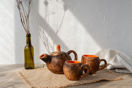 Aesthetic handcraft pottery tea set, brown clay teapot and muds on a wooden table, sun light shadows on white wall background, minimalist tea ceremony composition, wabi sabi conceptの写真素材