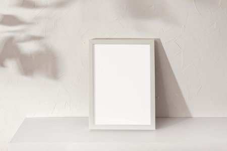 Blank picture frame with mockup copy space standing on white shelf, floral sun light shadows on a white concrete wall background, minimalist sustainable branding or interior design templateの写真素材
