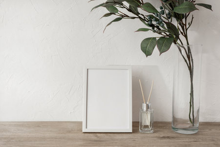 Blank photo frame, green leaves on vase, home fragrance on a beige wooden tabletop, white plaster wall background, minimalist brand or interior design template, mockup with copy spaceの写真素材