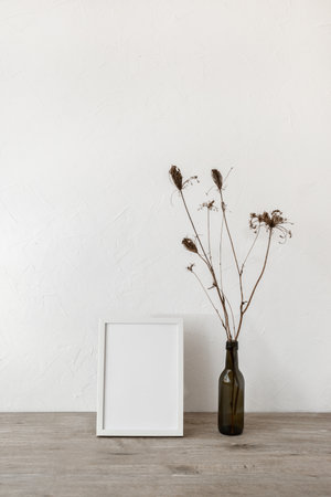 Empty picture frame and vase with flowers on a neutral beige wooden tabletop, white wall background,...
