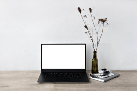 Blank laptop screen mockup, minimalist home office workspace, vase with flowers, stationery and opened laptop on a neutral beige wooden tabletop, white wall background, template with copy spaceの写真素材