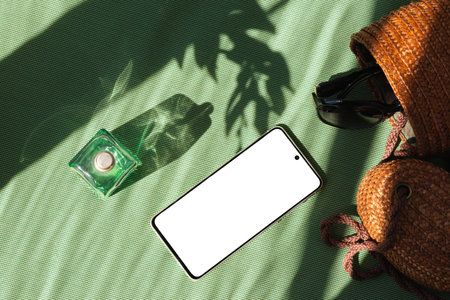 Summer fashion background with mobile phone and woman accessories on a green textile with sun light shadow, blank screen mock up with copy spaceの写真素材