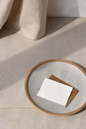 Blank paper card and envelope on ceramic gray plate, on beige stone table with sunlight shadow, minimalist business brand template, invitation or greeting card mockup, copy spaceの写真素材
