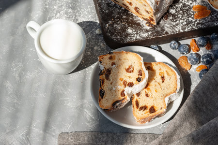 Sweet fruit cake, stollen with raisins and blueberries, sliced on plate, cup of milk, authentic Christmas morning breakfast with sunlight shadows on a gray background, copy spaceの写真素材