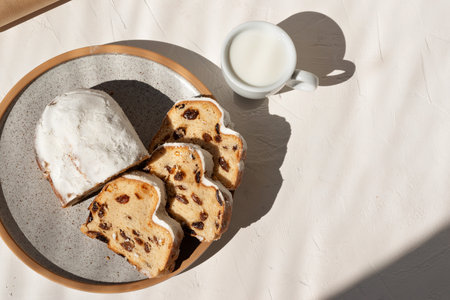 Traditional Christmas stollen sliced on a plate, cup of milk on beige background, sweet fruit cake with raisins and shugar powder, lifestyle morning breakfast flatlay with sunlight shadows, copy spaceの写真素材
