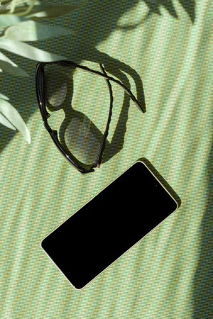 Minimal summer fashion concept, sunglasses and mobile phone on bright green background with abstract sun light shadow, copy spaceの写真素材