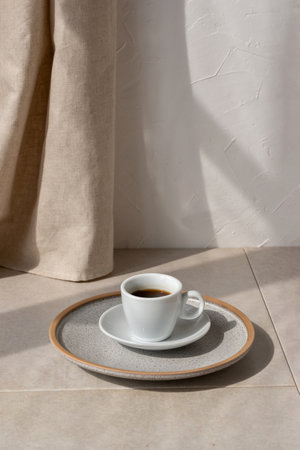 Lifestyle morning coffee concept, coffee cup in plate on beige table, warm neutral interior, beige linen curtain and empty white wall with sunlight shadow and copy spaceの写真素材