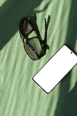 Summer fashion stylish backdrop, mobile phone with blank screen mock up, sunglasses on a green textile background with aesthetic abstract sun light shadows, minimal business brand template, copy spaceの写真素材