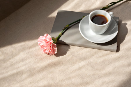 Minimalist aesthetic summer still life, coffee cup, notebook and carnation flower on a beige table with abstract lifestyle sunlight shadowsの写真素材