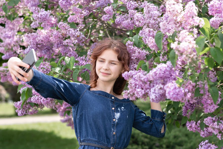Pretty teenager girl in denim dress taking selfie in a spring blooming park among lilac flowers, with eyes closedの写真素材