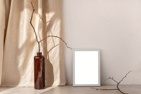 Empty picture frame mockup standing on a stone tabletop, brown vase with wooden branch, sunlight shadows on linen curtain and neutral beige wall background, minimalist design template, copy spaceの写真素材