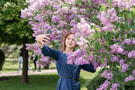 Happy young teenager girl in denim dress looking at screen of mobile phone and taking a selfie in lilac blooming garden, girl taking self portrait photo in flowers for social media or blogの写真素材