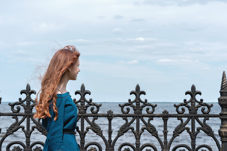 Cure serene teenager girl with red hair looking at sea and sky nature landscape, enjoying sunny weather and wind breeze outdoor on the embankmentの写真素材