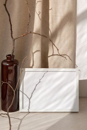 Blank picture frame with mockup copy space on a beige table, vase with branches, abstract sun light shadows on linen curtain background, minimalist sustainable branding or interior design templateの写真素材