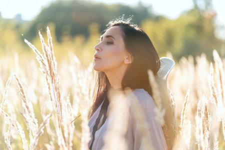 Beautiful lifestyle middle age woman holding meadow spikelets in a hand and blowing on it, pollen and dust in air, blurred defocused portrait with backlighting haze, allergic season conceptの写真素材