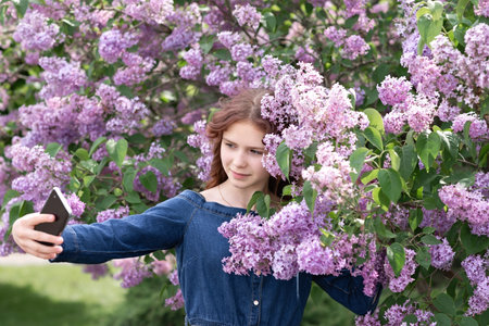 Beautiful teenager girl with red hair standing under blooming lilac trees in garden or park, taking...