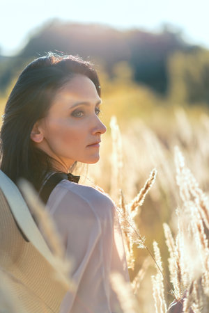 Beautiful middle age woman in meadow grass on summer sunset, walking in a nature outdoor, serenity, calm and relaxation concept, blurred defocused portrait with backlighting hazeの写真素材