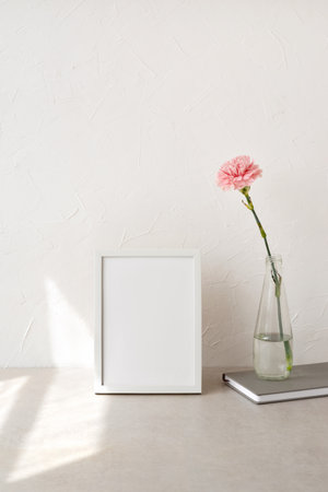Empty white picture frame, art poster mockup, vase with carnation flower, notebook on a neutral beige table and concrete wall background, aesthetic minimalist home workspace with sunlight patternの写真素材