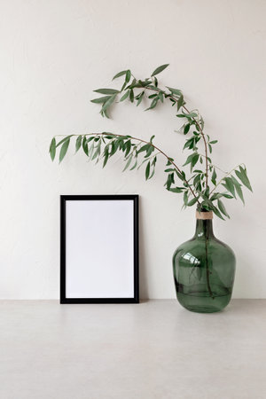 Empty black picture frame mockup, green glass jar with tree branches on neutral beige table, concrete wall background. Modern interior decoration, aesthetic minimal art poster template, copy spaceの写真素材