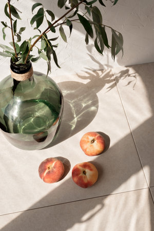 Aesthetic lifestyle still life with peaches, green glass jar with plants and sun light shadows on a beige linen tablecloth background. Summer slow living conceptの写真素材
