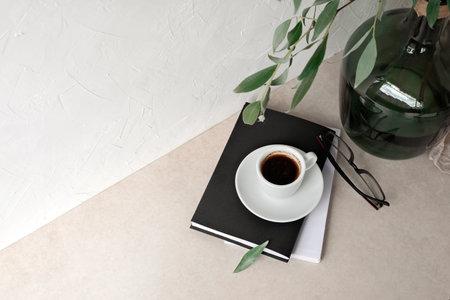 Coffee cup, books, glasses, green glass bottle with tree branch on beige tabletop, empty wall background. Minimalist business branding template, home office workspace flatlayの写真素材
