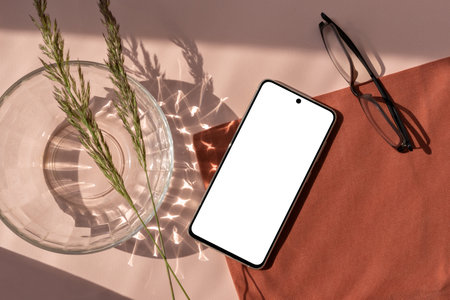 Empty mobile phone screen mockup, glasses, vase and meadow grass on a light pink and terracotta background with abstract sunlight shadow and crystal reflections, minimal business brand templateの写真素材