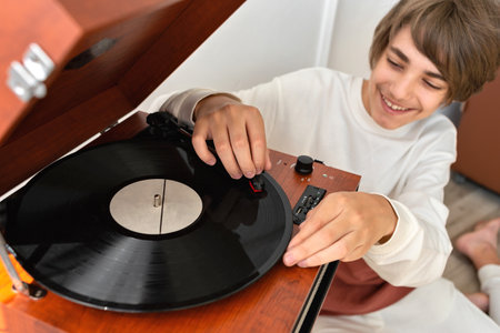 Happy smiling handsome teenager boy turning on retro vinyl player, brown wooden turntable and listening musicの写真素材