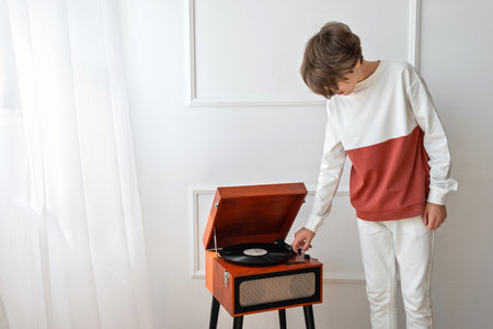 Teenager boy turning on retro vinyl player, brown wooden turntable, listening music on a white room backgroundの写真素材