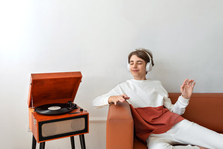 Happy beautiful teenager boy sitting on a brown sofa, listening vinyl record on retro wooden turntable, smiling, enjoying music, having fun. Empty white wall backgroundの写真素材