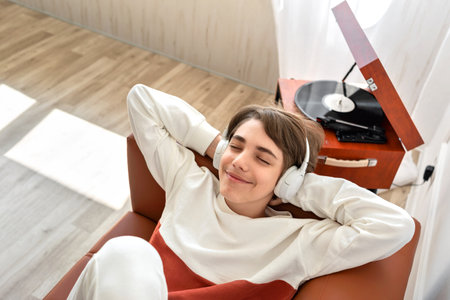 Handsome teenager boy wearing earphones listening vinyl records on retro wooden turntable, laying on a brown sofa and smiling, enjoy music soundの写真素材