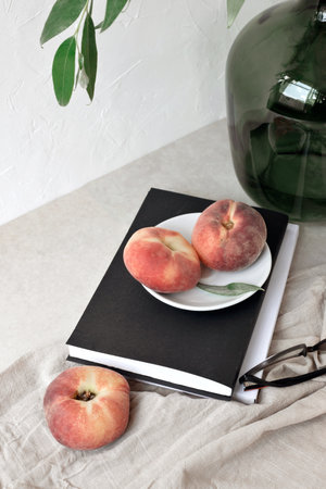 Aesthetic summer still life, ripe peach fruit, book, glasses, green vase with tree branch on linen tablecloth, empty white wall backgroundの写真素材