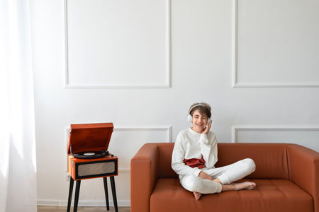 Beautiful happy teenager boy sitting on brown sofa, listening vinyl record on a retro wooden turntable, smiling, enjoying music, having fun. Empty white wall backgroundの写真素材