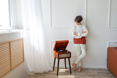 Young teenager boy standing near retro wooden vinyl turntable, looking on player and listening music indoor, in a white minimalist room interiorの写真素材