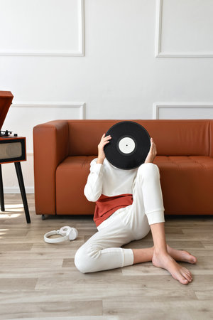 Teenager boy sitting on a floor near brown sofa and wooden turntable, holding vinyl record in hands, hiding face behind vinyl record. Retro vinyl music trend aesthetic.の写真素材