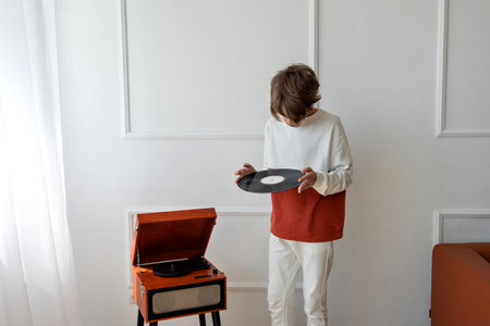 Teenager boy standing near brown wooden turntable, looking on a retro vinyl record, that he holding in hands. Home interior, white room backgroundの写真素材