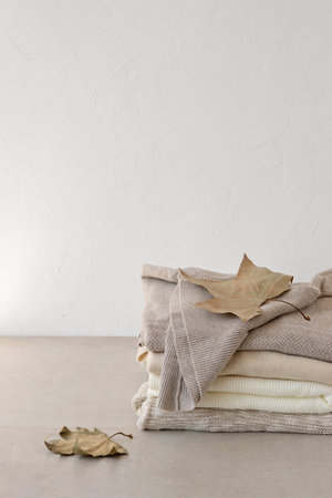 Neutral beige pastel wool, cashmere and cotton sweaters stack with dried autumn leaves on table, on empty white wall background. Fall warm knitwear, aesthetic minimalist fashion wardrobe.の写真素材