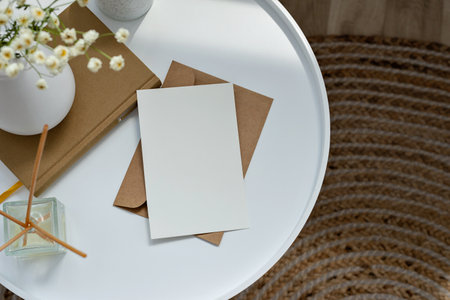 Aesthetic boho still life, empty paper card mockup, envelope, book, vase with flowers on white table, brown jute rug backgroundの写真素材