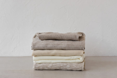 Minimalist neutral pastel beige wool, cashmere knitted sweaters stack on beige table on empty white wall background. Autumn, fall fashion woman clothes, knitwear wardrobeの写真素材