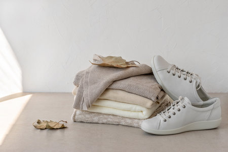 Fashion minimalist autumn clothes wardrobe in neutral beige colors. Beige, creamy, taupe wool knitted sweaters stack, white sneakers, brown fall leaves on table, on white empty wall backgroundの写真素材