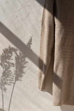 Aesthetic minimalist sustainable woman fashion clothes concept. Light brown, neutral beige wool sweater hanging on white empty wall background with lifestyle floral sunlight shadow.の写真素材