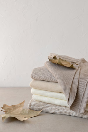 Early autumn fashion concept. Minimalist neutral beige knitted sweaters stack, fall leaves, wardrobe for september. Empty white wall background, copy space.の写真素材