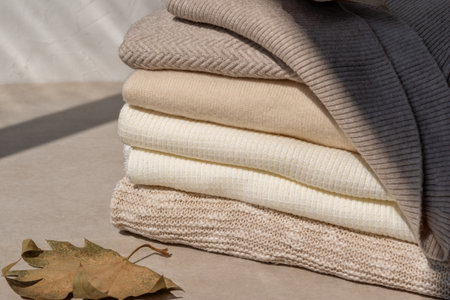 Stylish casual neutral nude color knitwear, autumn elegant woman clothes wardrobe. Beige knitted woolen jersey sweaters stack on table, fall leaf, sunlight shadows.の写真素材